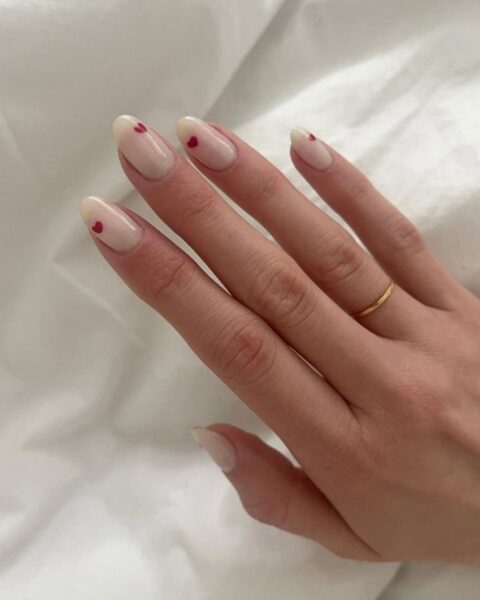 Subtelny manicure na długich, owalnych paznokciach. Baza to mleczny, półprzezroczysty nude w odcieniu ciepłego beżu, idealny dla estetyki clean look. Na każdym paznokciu znajduje się jeden minimalistyczny, czerwony detal w kształcie serca, umieszczony centralnie przy nasadzie, co nadaje stylizacji romantyczny i jednocześnie wyrafinowany charakter. Dłoń zdobi klasyczny, cienki, złoty pierścionek. Idealny przykład inspirującego mani dla kobiet ceniących dyskretną elegancję.