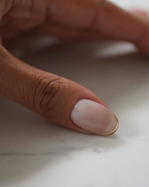 Wyrafinowany manicure na owalnym paznokciu. Kolor to mleczny, półprzezroczysty róż (sheer milk-pink), który idealnie imituje zdrową płytkę paznokcia. Charakterystycznym detalem jest złoty pasek (lub złoty mikro-french), delikatnie obrysowujący krawędź paznokcia. Ten minimalistyczny, luksusowy i inspirujący detal nadaje stylizacji wyrafinowania i jest idealnym wyborem dla kobiet z klasą, poszukujących eleganckich trendów w urodzie (np. manicure ślubny). Ujęcie w zbliżeniu na białym, marmurowym tle.