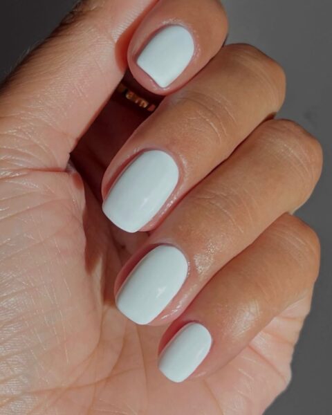Białe paznokcie o kremowym, kryjącym wykończeniu, idealne na zimowy manicure. Krótko spiłowane, zadbane paznokcie z błyszczącym topem, które symbolizują czystą elegancję i minimalizm w urodzie. Jasny, rozświetlający odcień bieli doskonale kontrastuje z karnacją, dodając stylizacji luksusowego, ponadczasowego charakteru.