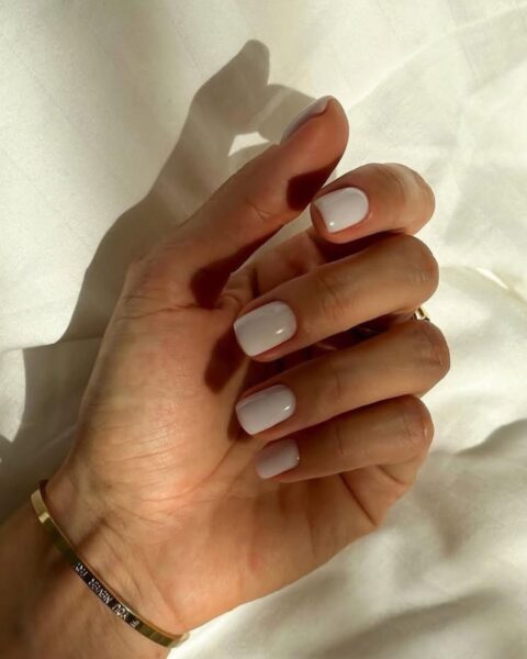 Subtelny, mlecznobiały manicure w stylu clean look lub nude, idealny dla kobiet ceniących ponadczasową elegancję. Krótkie, kwadratowe paznokcie pokryte lakierem o chłodnym odcieniu bieli, który pięknie harmonizuje z naturalną karnacją. Na nadgarstku widać minimalistyczną, złotą bransoletkę z grawerem, podkreślającą luksusowy i inspirujący detal stylizacji.