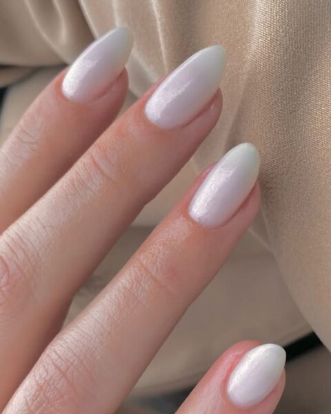 Perłowy, opalizujący manicure w odcieniu mlecznej bieli z delikatnym, różowym błyskiem. Paznokcie o modnym, migdałowym kształcie i średniej długości, pokryte lakierem dającym efekt glamour (lub "glazed donut nails"). Ten świetlisty, wyrafinowany kolor dodaje dłoniom eterycznej elegancji i jest inspirującym wyborem dla kobiet, które cenią subtelny luksus. Tło to lśniący, beżowy materiał.