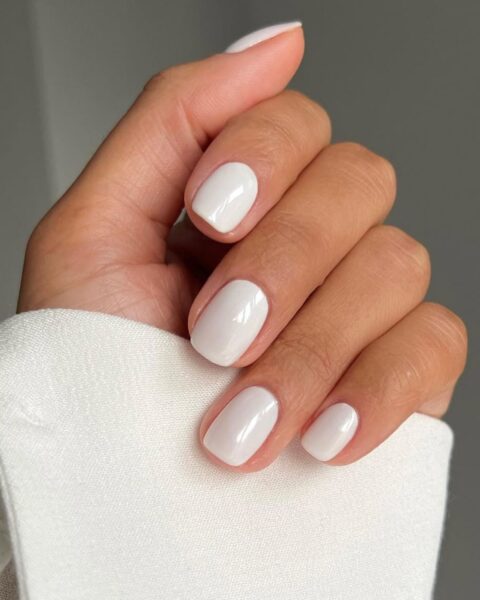 Lśniący, perłowy manicure w odcieniu mlecznej bieli na krótkich, kwadratowych paznokciach. Kolor charakteryzuje się subtelnym, satynowym połyskiem (efekt "perfect pearl"), który nadaje paznokciom wyrafinowanego i luksusowego charakteru. Ten czysty, wyjątkowy odcień bieli symbolizuje minimalistyczną elegancję. Ujęcie eksponuje zadbaną dłoń i fragment śnieżnobiałej, marszczonej koszuli.
