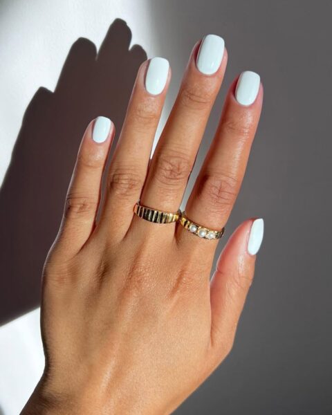 Wyrazisty, pastelowy manicure w odcieniu miętowej bieli (lub lodowej mięty), który rozjaśnia dłoń i dodaje jej świeżości. Ten chłodny, subtelny kolor doskonale komponuje się z opaloną skórą, nadając paznokciom o owalnym kształcie nowoczesnego, a jednocześnie wyrafinowanego wyglądu. Dłoń zdobią dwa złote pierścionki – jeden o fakturowanej powierzchni, drugi z perłami – co podkreśla luksusowy i inspirujący charakter stylizacji. Cień na tle dodaje zdjęciu głębi i artystycznego wyrazu.