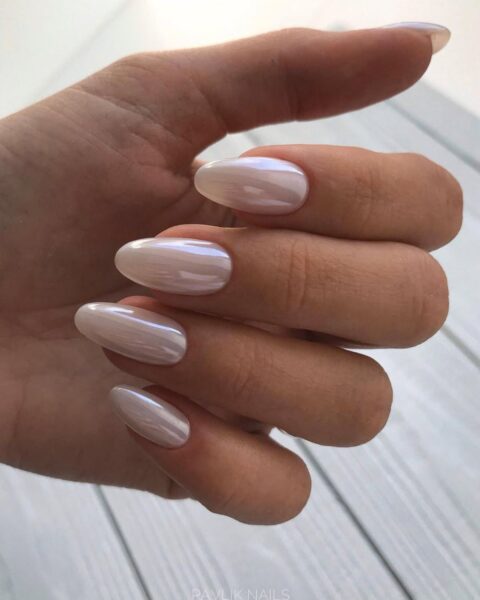 Eteryczny manicure o migdałowym kształcie paznokci, pokryty perłowym lakierem w odcieniu rozbielonego beżu (lub nude z efektem lustra). Charakterystyczny lustrzany połysk i delikatna opalizacja nadają stylizacji wyrafinowanego i modnego charakteru "glazed donut nails". Ten świetlisty, elegancki kolor jest idealny dla kobiet ceniących subtelny luksus w urodzie. Ujęcie eksponuje długie, zadbane paznokcie w naturalnym świetle.