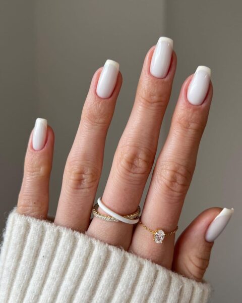 Białe paznokcie na jesień w mlecznym odcieniu (tzw. milky white). Manicure z delikatną, subtelną końcówką (w stylu mikro-french lub baby boomer), która dodaje stylizacji wyrafinowania i klasy. Krótkie, kwadratowe paznokcie doskonale komponują się z ciepłym, inspirującym detalem: białym, wełnianym swetrem i minimalistycznymi złotymi pierścionkami (w tym pierścionek z białą emalią i delikatny łańcuszek z diamentem). To ucieleśnienie cichego luksusu w urodzie.