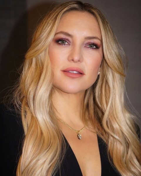 Bliska fotografia Kate Hudson z długimi, warstwowymi, miodowymi falami blond, ułożonymi z przedziałkiem na środku. Aktorka ma intensywny makijaż oczu z cieniem w kolorze burgundowym i usta podkreślone błyszczącą, różową szminką. Nosi złoty, delikatny naszyjnik z wisiorkiem.