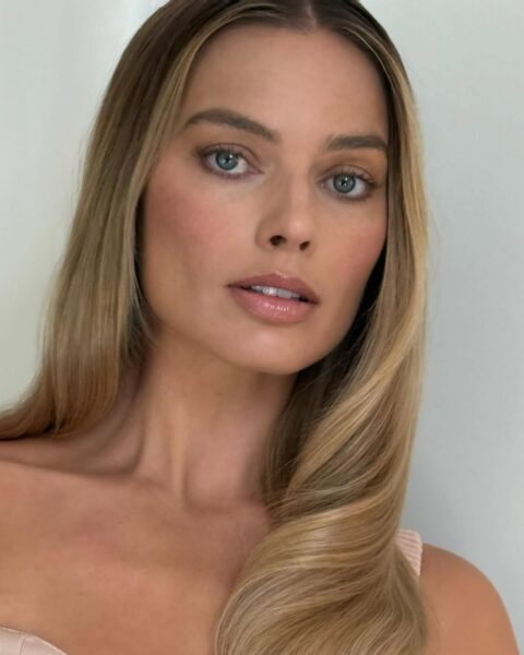 Bliska fotografia Margot Robbie z długimi, jasnymi włosami blond ułożonymi w miękkie, duże fale i przedziałkiem na boku. Aktorka ma subtelny, naturalny makijaż, podkreślający jej oczy i kości policzkowe brzoskwiniowym różem. Ma na sobie jasnoróżowy top na ramiączkach.