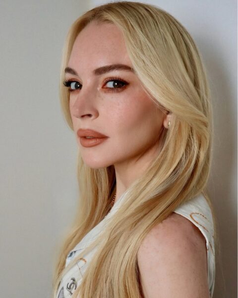 Bliska fotografia Lindsay Lohan z długimi, platynowo-blond włosami, zaczesanymi na bok. Aktorka ma wyrazisty makijaż z matową pomadką w odcieniu terakoty i smokey eye w ciepłych, miedzianych tonach. Ma na sobie białą koszulę bez rękawów z delikatnym wzorem.
