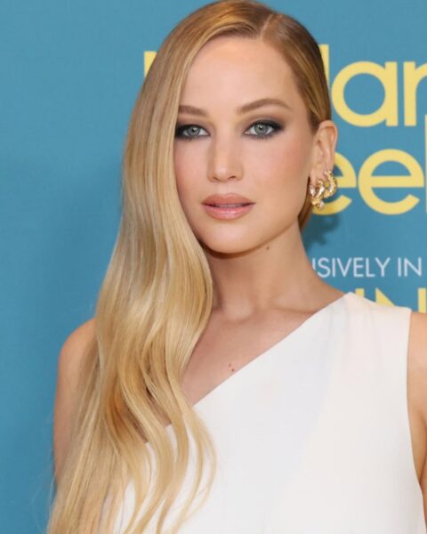 Bliska fotografia Jennifer Lawrence w białej sukni na jedno ramię. Aktorka ma długie, ciepłe blond włosy, ułożone w głęboki, boczny przedziałek z miękkimi falami, oraz wyrazisty, przydymiony makijaż oczu (smokey eye) i błyszczącą, cielistą szminkę. Nosi złote, rzeźbione kolczyki.
