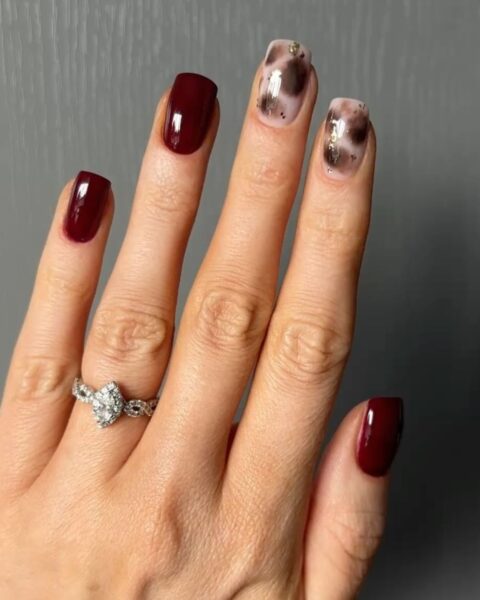 Modny jesienny manicure typu mix & match, łączący klasyczne burgundowe paznokcie o kwadratowym kształcie z dwoma paznokciami ozdobionymi abstrakcyjnym, marmurkowym wzorem w odcieniach brązu, beżu i bieli ze złotymi akcentami (imitacja liści szlagmetalu). Głęboki burgund jest błyszczący i elegancki. Na palcu serdecznym widoczny jest pierścionek zaręczynowy z dużą łezkową cyrkonią i zdobioną obrączką.