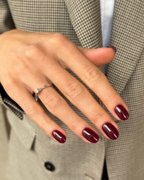 Elegancki, jesienny manicure z klasycznymi, głębokimi burgundowymi paznokciami o owalnym kształcie i wysokim połysku. Dłoń z paznokciami ułożona na marynarce w szaro-brązową kratę (pepita/księcia Walii), co podkreśla szykowny i profesjonalny charakter stylizacji. Na palcu serdecznym widoczny srebrny pierścionek z drobnym diamentem lub cyrkonią. Idealny wybór dla kobiety z klasą.