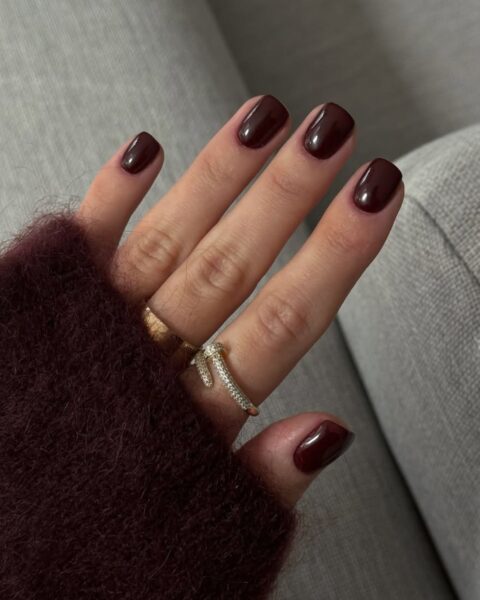 Głębokie burgundowe paznokcie o kwadratowym kształcie i wysokim połysku, idealne na jesień. Manicure jest w tonacji dopasowanej do ciepłego, włochatego swetra (bordowy lub ciemnoczerwony). Dłoń zdobią złote pierścionki, w tym jeden z motywem węża wysadzany cyrkoniami.