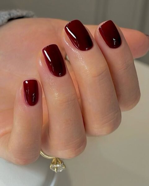 Piękny jesienny manicure – błyszczące, burgundowe paznokcie o kwadratowym/zaokrąglonym kształcie, idealnie pokryte głębokim, winnym odcieniem. Manicure wykonany metodą BIAB (Builder in a Bottle), który wzmacnia naturalną płytkę. Paznokcie w kolorze burgundy prezentują się elegancko, a dłoń zdobi dyskretny złoty pierścionek z przezroczystym kamieniem.
