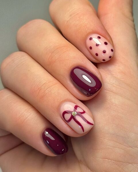 Modny jesienny manicure łączący klasyczne burgundowe paznokcie z delikatnym, cielistym tłem. Paznokcie o owalnym kształcie z błyszczącym wykończeniem. Wybrane płytki zdobią wzory: kokardka z cyrkonią oraz delikatne burgundowe kropki na jasnym, mlecznoróżowym tle. Eleganckie i dziewczęce zdobienie, idealne dla kobiet ceniących klasę i subtelny detal.