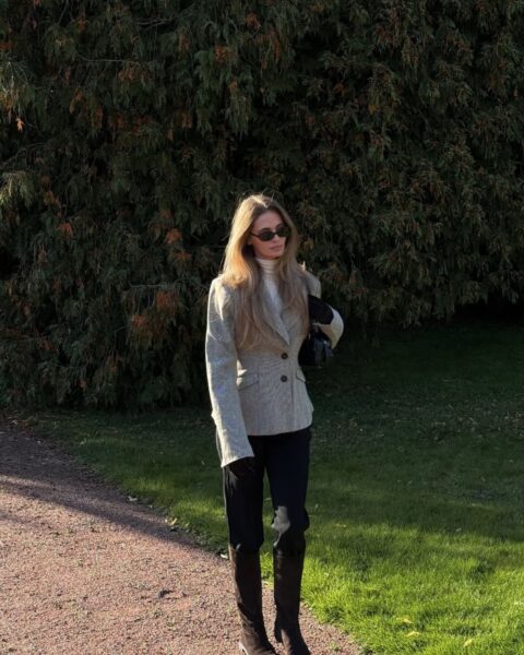Kobieta w eleganckiej, jesiennej stylizacji w stylu smart casual. Ma na sobie tweedową lub wełnianą marynarkę w odcieniach beżu lub szarości (wzór jodełka/pepitka), pod którą widać biały golf. Do tego czarne spodnie i czarne kozaki z cholewką do kolana. Modelka stoi na żwirowej ścieżce w parku, w tle duże, zielone krzewy. Inspiracja: jak nosić marynarkę w kratę i biały golf, klasyka jesiennej mody.