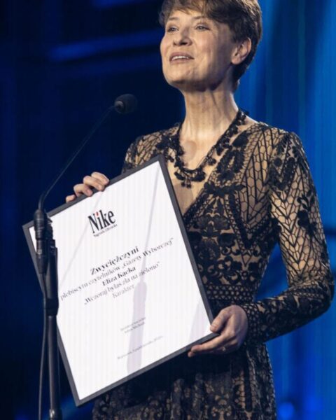Eliza Kącka na scenie, w czarnej koronkowej sukience, trzymająca dyplom potwierdzający zdobycie nagrody literackiej Nike Czytelników za książkę "Wczoraj byłaś dla mnie najpiękniejsza".