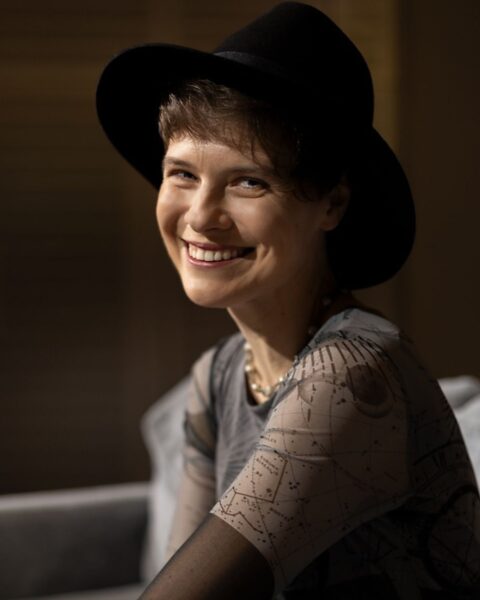 Eliza Kącka uśmiecha się na portrecie, ubrana w czarny kapelusz typu fedora, półprzezroczystą bluzkę z motywem konstelacji i delikatny sznur pereł.