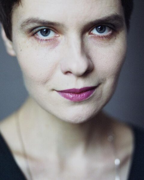 Eliza Kącka na zbliżeniu (close-up), z intensywnym spojrzeniem i delikatnym uśmiechem, w minimalistycznej stylizacji z różową szminką i subtelnym naszyjnikiem.