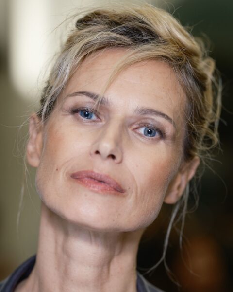 Zdjęcie z bliska przedstawia kobietę z lekko zmierzwionym, eleganckim upięciem włosów w kolorze blond. Pojedyncze kosmyki luźno opadają na jej czoło. Kobieta ma na sobie szarą marynarkę, a jej makijaż jest naturalny i subtelny. Fryzura i stylizacja są dobrym przykładem klasycznej, ponadczasowej elegancji.