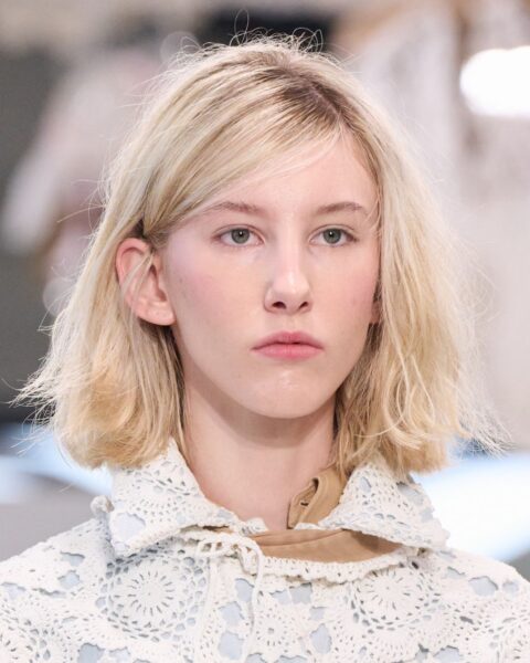 Zdjęcie przedstawia młodą kobietę z krótką, blond fryzurą typu bob. Włosy są naturalnie pofalowane, z delikatną grzywką opadającą na czoło i zaczesanym za ucho kosmykiem. Kobieta ma na sobie jasną, koronkową bluzkę. Fryzura jest swobodna i dziewczęca.
