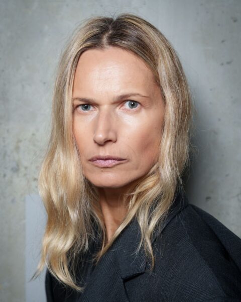 Zdjęcie portretowe przedstawia kobietę z długimi, blond włosami z przedziałkiem na środku. Włosy są stylizowane na delikatne, luźne fale i sprawiają wrażenie naturalnie pofalowanych. Kobieta ma na sobie elegancką marynarkę w ciemnym kolorze. Całość prezentuje klasyczną elegancję i prosty, ale wyrafinowany styl.