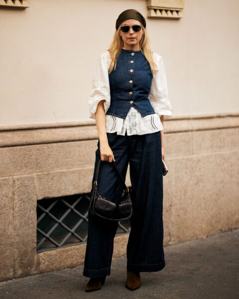 Stylowa kobieta w modnej, inspirowanej stylem retro stylizacji street style w Mediolanie. Ma na sobie białą koszulę z bufiastymi rękawami i koronkowymi detalami, na którą nałożona jest dopasowana kamizelka z ciemnego denimu z ozdobnymi guzikami. Dół to bardzo szerokie jeansy typu palazzo. Całość uzupełnia opaska na włosach, mała czarna torebka na ramię i brązowe botki.