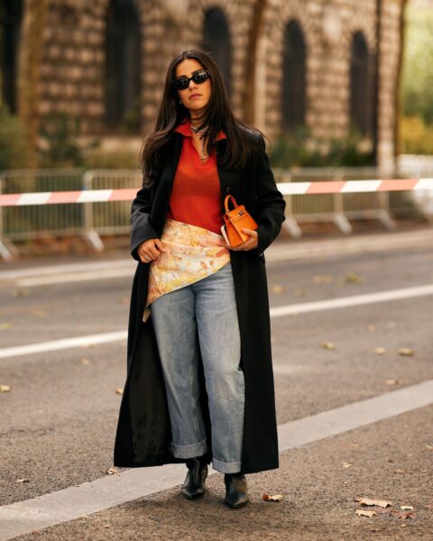 Stylowa kobieta w modnej stylizacji street style w Paryżu: czarny długi płaszcz, pomarańczowy sweter polo i luźne jeansy podwinięte nad czarnymi kowbojkami. Jak nosić kowbojki i jeansy, to połączenie nonszalancji i klasy, z dodatkiem pomarańczowej torebki i wzorzystej apaszki przewiązanej w talii.