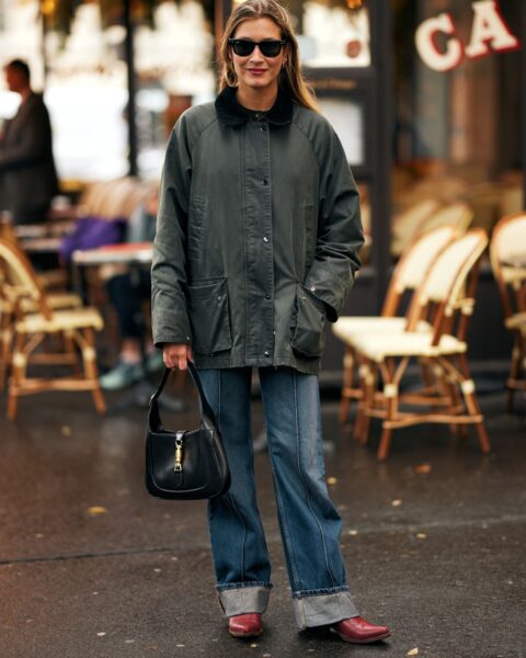 Elegancka stylizacja street style sfotografowana w Paryżu: kobieta w klasycznej, woskowanej kurtce w kolorze khaki z kołnierzem, szerokich jeansach z podwiniętymi nogawkami oraz burgundowych botkach. Akcesoria dodające szyku: czarna torebka-bagietka i prostokątne okulary przeciwsłoneczne.