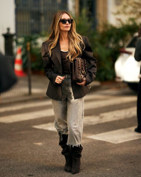 Elegancka stylizacja street style w odcieniach brązu i szarości. Kobieta ma na sobie czarną, dopasowaną marynarkę, luźne, sprane jeansy z surowym wykończeniem oraz czarne, zamszowe kowbojki z szeroką, marszczoną cholewką. Jak nosić kowbojki i jeansy z klasą: ten look uzupełnia duża, pikowana torebka na łańcuchu w kolorze czekoladowego brązu i prostokątne okulary przeciwsłoneczne.