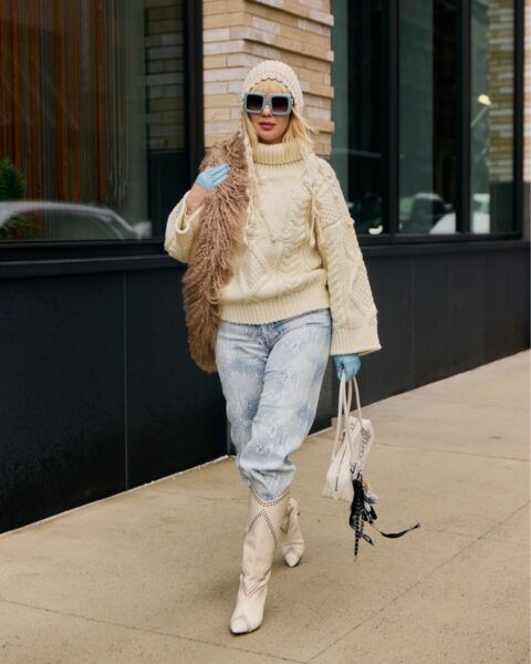 Efektowna stylizacja street style w odcieniach beżu i bieli: kobieta w kremowym swetrze z golfem o warkoczowym splocie, czapce beanie i jasnych jeansach. Całość uzupełnia beżowe futrzane boa, błękitne rękawiczki oraz białe, zdobione kowbojki i mała torebka. Szykowny look podkreślają duże, kwadratowe okulary przeciwsłoneczne z błękitnymi szkłami.
