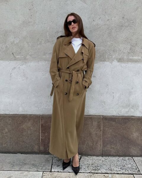 Kobieta w eleganckim, długim, beżowym trenczu z paskiem, czarnych szpilkach i białym T-shircie, stojąca przy dwukolorowej ścianie. Stylizacja pokazuje, jak stylizować trench na elegancki, miejski look.