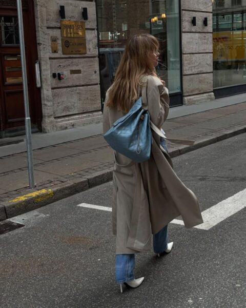 Kobieta idąca ulicą, sfotografowana od tyłu. Ma na sobie beżowy, długi trencz, proste jeansy, białe botki na obcasie oraz dużą, pikowaną torebkę w odcieniu błękitu, przewieszoną przez ramię.