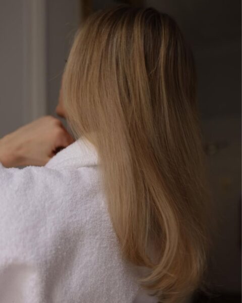 Zdjęcie od tyłu, skupiające się na długich, prostych i lśniących blond włosach, lekko podwiniętych na końcach. Kobieta ma na sobie biały szlafrok frotte i dotyka ręką ramienia. Ujęcie sugeruje moment pielęgnacji i zdrowy wygląd włosów.
