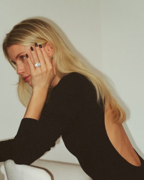 Kobieta o długich, prostych, platynowych włosach blond patrzy w bok, opierając głowę o dłoń. Ma na sobie elegancką, czarną sukienkę z głębokim wycięciem na plecach. Uwagę zwraca duży, wyrazisty pierścionek na palcu oraz ciemny manicure. Zdjęcie jest zrobione w ciepłym, nieco ziarnistym świetle, podkreślającym klasyczny, ale zmysłowy styl.