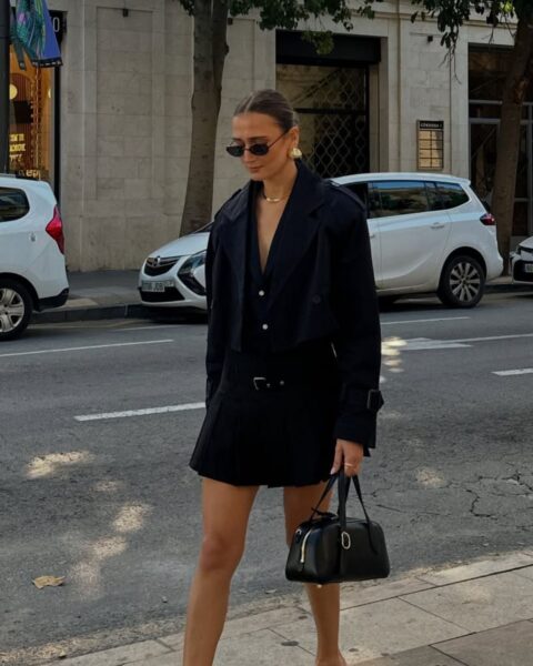 Kobieta idąca ulicą w monochromatycznej, granatowej stylizacji. Ma na sobie krótki, pudełkowy, sportowy żakiet (cropped jacket), mini spódniczkę w kratę (lub plisowaną mini) oraz czarne klapki na obcasie typu mule. Stylizację dopełnia czarna torebka-kuferek, owalne okulary przeciwsłoneczne i minimalistyczna złota biżuteria. Modne stylizacje z krótkimi żakietami w stylu 'preppy chic' na lato i jesień.