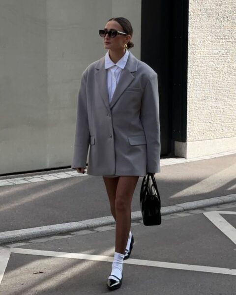 Kobieta w modnej, minimalistycznej stylizacji typu 'oversize'. Ma na sobie długi, luźny szary blezer (marynarkę) noszony jako sukienka, pod którym widać białą koszulę. Look dopełniają czarno-białe czółenka (mary-janes lub sling-back) w połączeniu z białymi skarpetkami. Całość uzupełniają kocie okulary przeciwsłoneczne, minimalistyczne złote kolczyki i czarna torebka. Inspiracja na modę w stylu 'preppy' i 'quiet luxury' na wiosnę.
