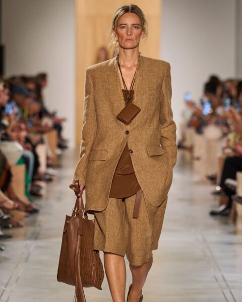 Modelka na wybiegu prezentuje stylowy, monochromatyczny zestaw w odcieniu ciepłego, postarzanego brązu (camel/cognac). Składa się on z oversizowej, wełnianej marynarki i pasujących do niej szortów typu bermudy o dłuższej nogawce. Pod spodem modelka ma brązową bluzkę. Całość uzupełniają: duża, brązowa skórzana torba z frędzlami, pasujące klapki na obcasie oraz naszyjnik z małą, skórzaną sakiewką. To klasyczny przykład minimalizmu w stylu quiet luxury, idealny dla kobiet poszukujących inspiracji do eleganckich, biznesowych stylizacji.