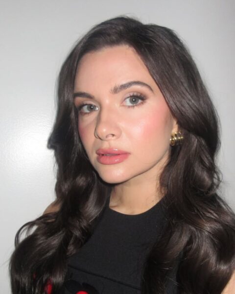 Zbliżenie na aktorkę Katie Stevens z gęstymi, ciemnymi, pofalowanymi włosami. Ma na sobie czarną bluzkę lub sukienkę ozdobioną dużymi, czerwonymi aplikacjami w kształcie kół, co nadaje stylizacji wyrazisty, geometryczny charakter. Makijaż jest klasyczny i promienny: mocny, dziewczęcy róż na policzkach, delikatny smoky eye w odcieniach brązu, oraz błyszczące, cukierkowe usta w kolorze baby pink. Całość dopełniają eleganckie, złote kolczyki z motywem kulek, podkreślające stylizację w duchu glamour z nutą retro.