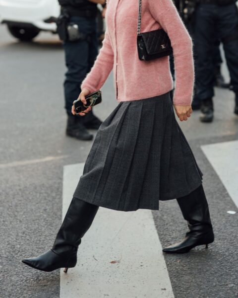 Stylowa kobieta w modnej jesiennej stylizacji street style, z mokrymi włosami zaczesanymi do tyłu, idzie przez pasy. Ma na sobie krótki, puszysty kardigan w kolorze pudrowego różu, szarą spódnicę midi plisowaną oraz czarne, skórzane kozaki z ostrym czubkiem i niskim obcasem. Na ramieniu wisi pikowana, czarna torebka na łańcuszku (Chanel), a jej twarz zasłaniają prostokątne okulary przeciwsłoneczne. W tle widać dwóch umundurowanych funkcjonariuszy policji i samochód.