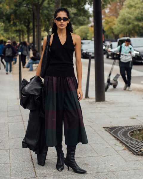 Młoda kobieta w eleganckiej, jesiennej stylizacji street style, stojąca na chodniku w miejskim otoczeniu (prawdopodobnie Paryż). Ma na sobie czarny, dzianinowy top typu halter z dekoltem w serek, szerokie spódnico-spodnie (kuloty) w szkocką kratę (burgundowo-zieloną) oraz czarne kozaki na niskim obcasie typu kitten heels. Stylizację dopełniają wąskie, czarne okulary przeciwsłoneczne. Na ramieniu wisi czarna, pleciona torebka, a w ręce trzyma złożoną czarną kurtkę (prawdopodobnie skórzaną). Włosy ma zaczesane do tyłu. W tle widać przechodniów, drzewa i ulicę.