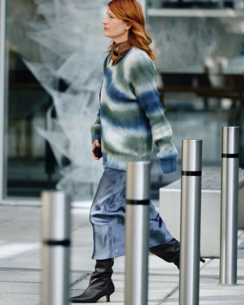 Kobieta w modnej jesiennej stylizacji street style idzie wzdłuż rzędu metalowych słupków, przed nowoczesnym budynkiem. Ma rude włosy, na szyi wiązaną apaszkę i duży, dzianinowy sweter w szerokie, rozmyte pasy w odcieniach zieleni, błękitu i szarości. Do tego dobrała jedwabną lub satynową spódnicę midi w kolorze chłodnego błękitu oraz czarne, skórzane kozaki na obcasie typu kitten heel.