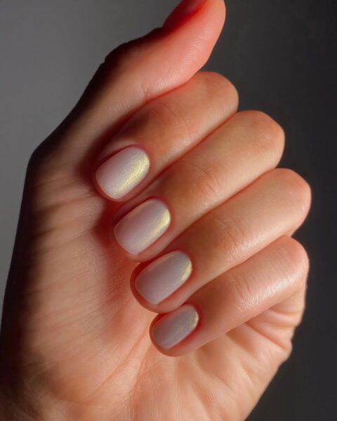 Modne, krótkie paznokcie w stylu "Glazed Donut Nails". Manicure w kolorze mlecznej bieli lub bardzo jasnego beżu z opalizującym, złotym lub szampańskim połyskiem (efekt chrome/perłowy). Lśniący lakier, prawdopodobnie odcień "Solar" marki Manucurist, który mieni się w świetle. Inspiracja eleganckim, nowoczesnym i minimalistycznym lookiem.