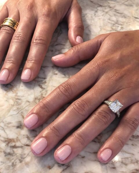 Krótkie paznokcie w kolorze nude - inspiracje. Manicure żelowy w delikatnym, mlecznym, jasnoróżowym odcieniu, idealny dla zadbanych dłoni i subtelnego looku. Na palcach widoczne złote pierścionki i duży pierścionek zaręczynowy z diamentem o szlifie szmaragdowym.