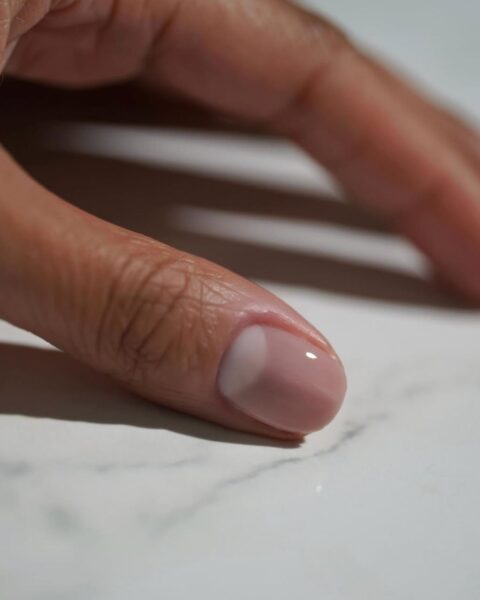 Manicure księżycowy (moon manicure) w nowoczesnym, mlecznym wydaniu. Paznokieć w kształcie migdała, pokryty dwoma odcieniami: beżowo-różowym (prawdopodobnie "Put it in Neutral" OPI) oraz mlecznobiałym półksiężycem u nasady (prawdopodobnie "Funny Bunny" OPI). Elegancka, minimalistyczna inspiracja dla naturalnych paznokci.