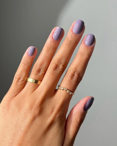 Modny kolor paznokci jesień/zima. Krótkie paznokcie pomalowane eleganckim, kryjącym lakierem w odcieniu lawendowej szarości lub zgaszonego fioletu (mauve/dusty lilac). Dłoń zdobią złote pierścionki z diamentami (w tym minimalistyczna, złota obrączka z elementami biżuterii w stylu Cartier Love). Inspiracja kolorem 'IT' według Toma Forda.
