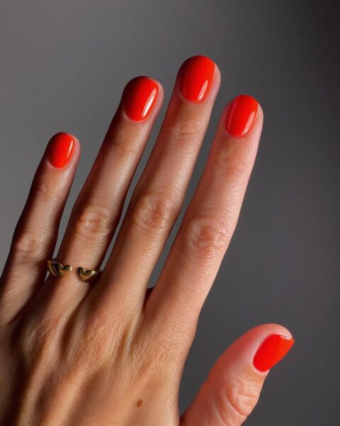 Krótkie paznokcie - inspiracje: Manicure w intensywnym, ognistym odcieniu czerwieni (prawdopodobnie lakier 'Lava' lub 'Red Cherry'). Kolor idealnie nasycony i lśniący. Dodatkowy akcent w postaci nowoczesnego moon manicure (manicure księżycowy) na kciuku, wykonanego w dwóch odcieniach czerwieni i bieli. Dłoń zdobi minimalistyczny, złoty pierścionek z motywem serca.