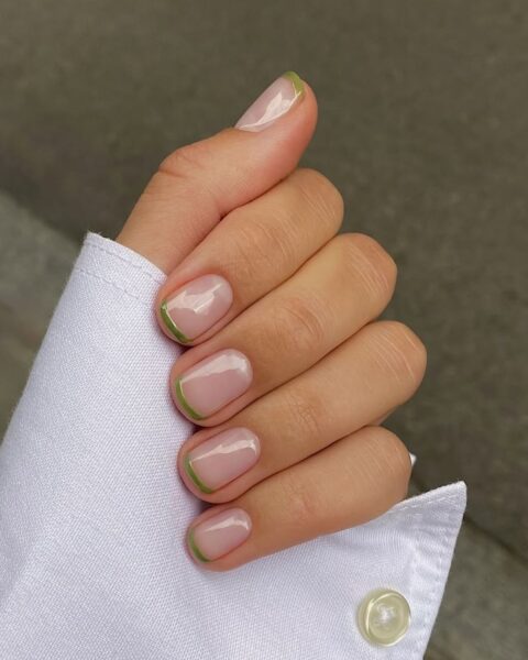 Nowoczesny French manicure w oliwkowej zieleni (olive french tips). Krótkie paznokcie o zaokrąglonym kształcie z minimalistyczną, cienką linią na końcówce paznokcia w kolorze ciemnej, zgaszonej zieleni (prawdopodobnie odcień 'Tuileries'). Baza paznokcia jest naturalna i mleczna. Elegancki, minimalistyczny trend manicure idealny dla kobiet z klasą. Dłoń spoczywa na mankiecie białej koszuli.