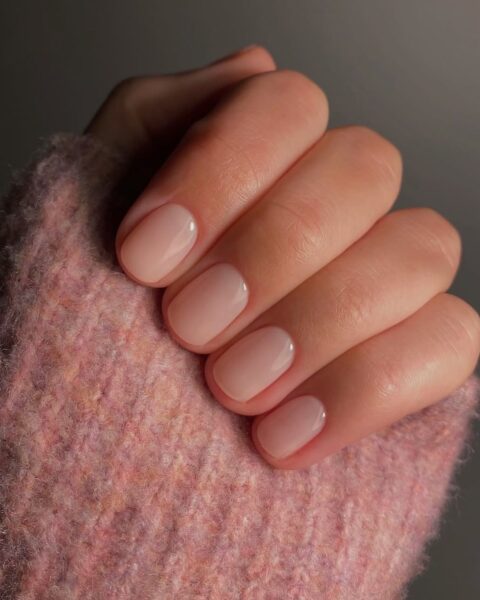 Subtelny manicure w stylu 'clean girl aesthetic' lub 'lip gloss nails'. Krótkie paznokcie w delikatnym, mlecznym odcieniu jasnego różu (prawdopodobnie "Sweet Candy Breath" Bio Sculpture), z lśniącym, żelowym wykończeniem. Minimalistyczny, elegancki i bardzo zadbany wygląd dłoni. W tle widoczny ciepły, różowy, wełniany kardigan, idealnie pasujący do paznokci.