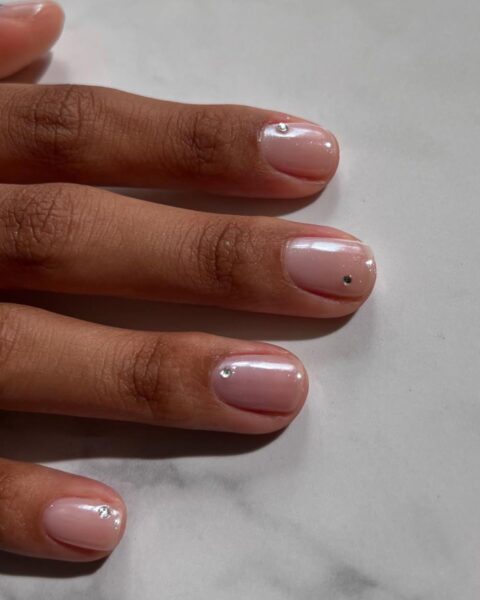 Krótkie paznokcie - inspiracje: "Naked nails" lub "glam nude" manicure. Paznokcie pokryte mlecznym, jasnoróżowym lakierem (prawdopodobnie "Bubble Bath" OPI) z subtelnym, perłowym efektem chrome. Zdobienie polega na umieszczeniu pojedynczej, minimalistycznej cyrkonii (kryształka Swarovskiego) blisko nasady paznokcia. Elegancki, dyskretny i uniwersalny manicure, idealny na każdą okazję.