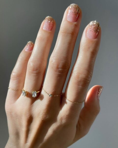 Świąteczny manicure w stylu glitter fade (gradient z brokatem) lub szampańskiego ombre. Paznokcie o naturalnym, owalnym kształcie z delikatną, różową bazą nude przechodzącą w złoty, lśniący brokat przy końcówkach. Ujęcie dłoni jest podkreślone przez minimalistyczną, złotą biżuterię – delikatne pierścionki łańcuszki z pojedynczymi brylantami lub kryształkami, stanowiące inspirację dla kobiet ceniących dyskretny luksus i blask idealny na eleganckie okazje.